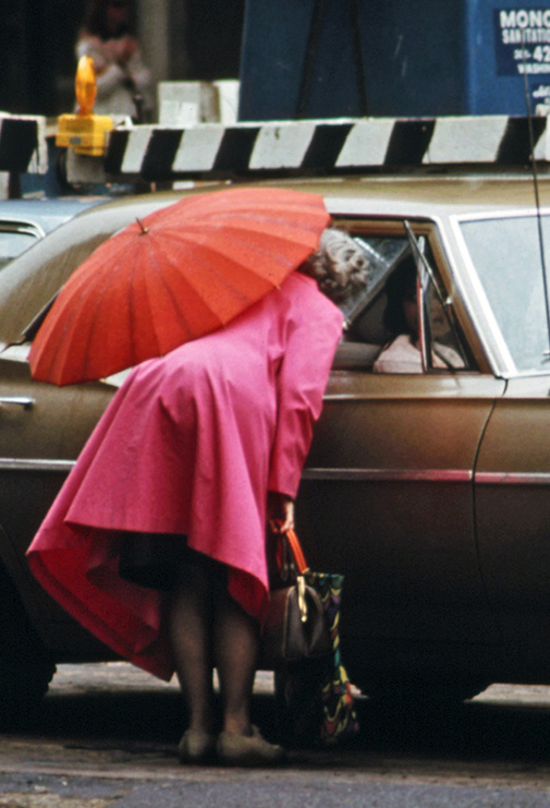 A woman in a pink coat holds a red umbrella while she bends down to talk to someone through a car window. The image is from the 70s and looks like a older archival photograph.